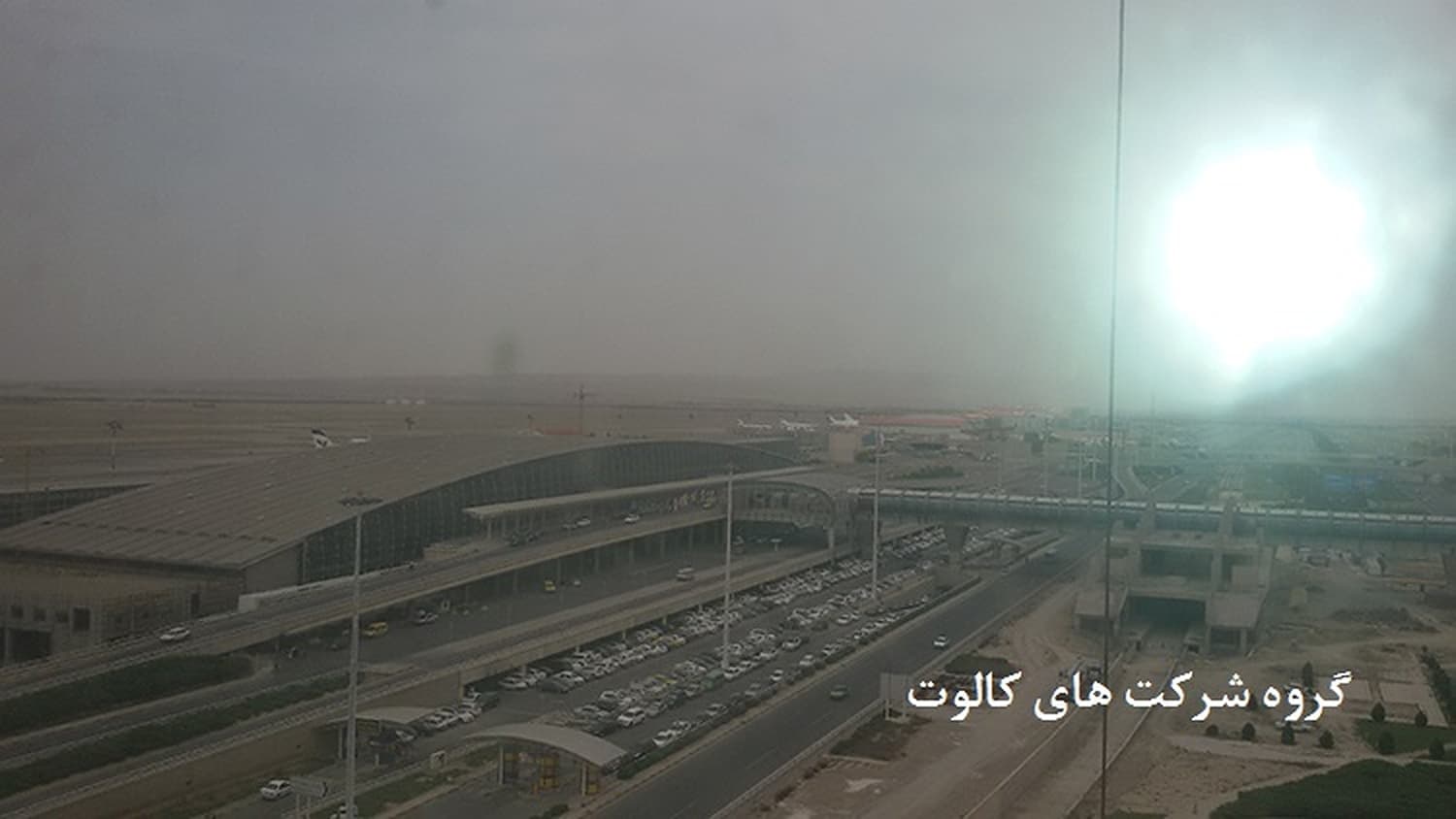ساختمان ترمینال فرودگاه با سقف قوسی فلزی و پارکینگ خودروها در نور مقابل خورشید