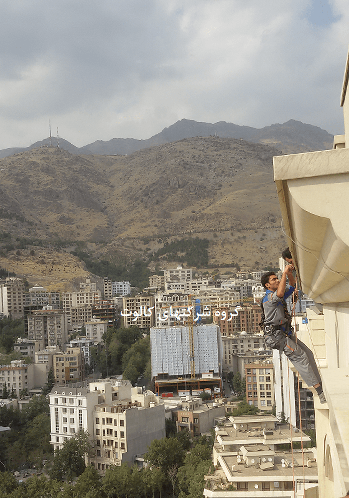 تکنسین با طناب و تجهیزات کار در ارتفاع در حال نصب هادی نزولی روی نمای ساختمان بلند، با شهر و کوه در پس‌زمینه