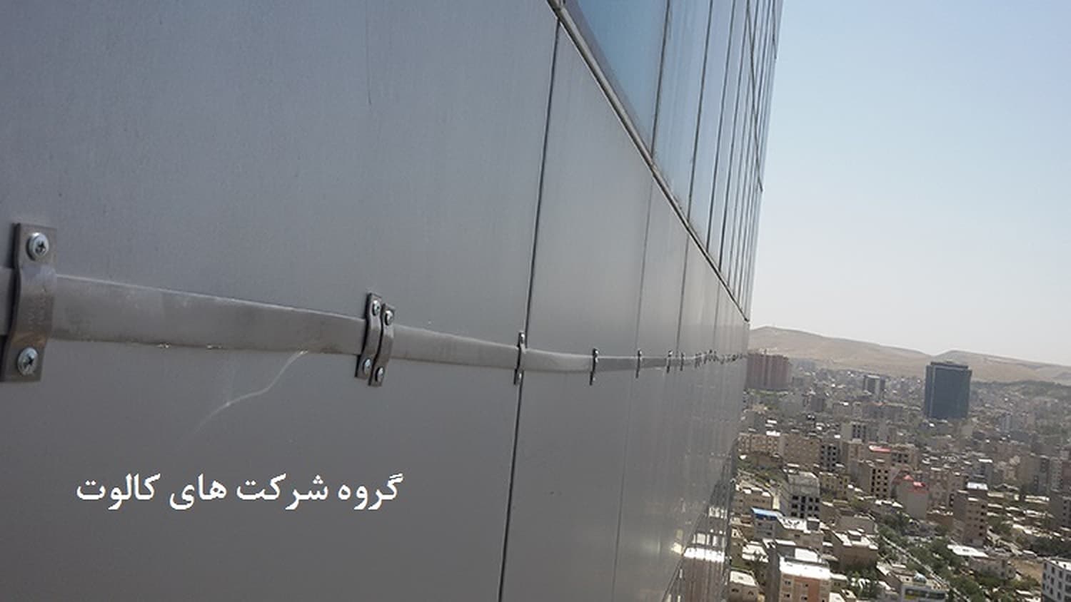 هادی نزولی با بست‌های نگهدارنده روی نمای فلزی ساختمان بلند، با نمای شهر در پس‌زمینه
